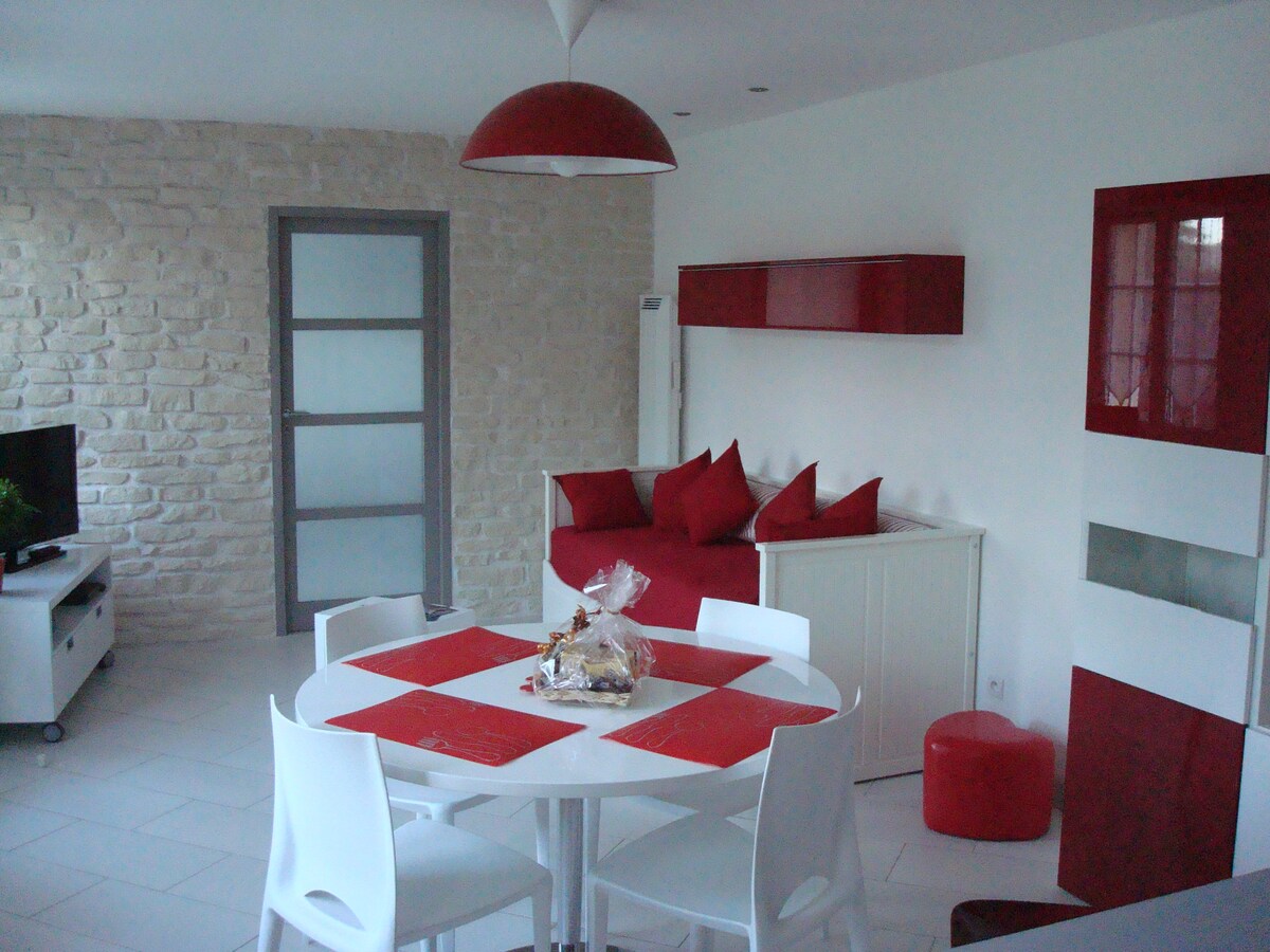 A modern living space features light-colored stone walls and a bright red accent decor. A round dining table with white chairs is centered in the room, surrounded by an inviting seating area with red cushions. A television is positioned against the wall.