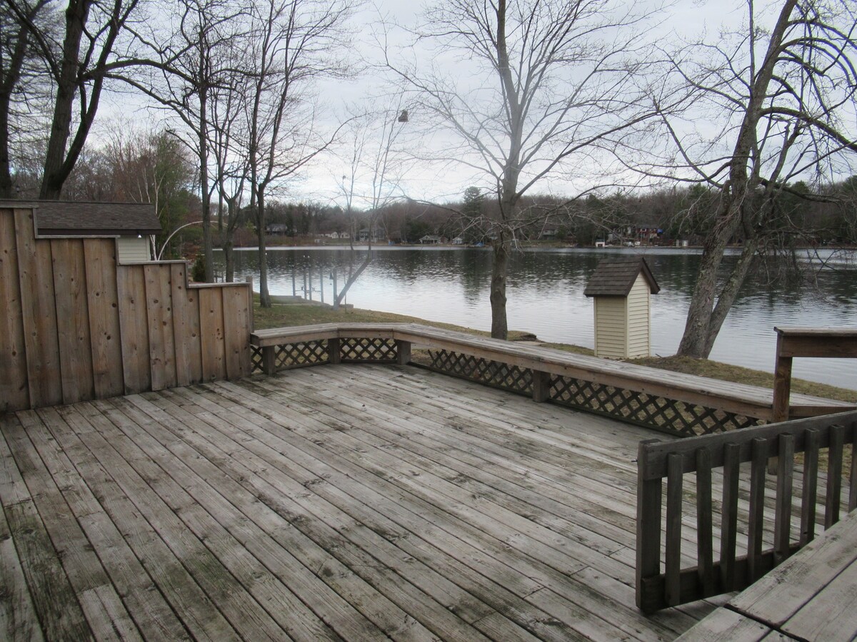 A spacious wooden deck extends towards the lake, bordered by low wooden railings. Trees provide a natural backdrop, while the water reflects the overcast sky. Nearby structures can be seen across the lake, contributing to a serene lakeside environment.