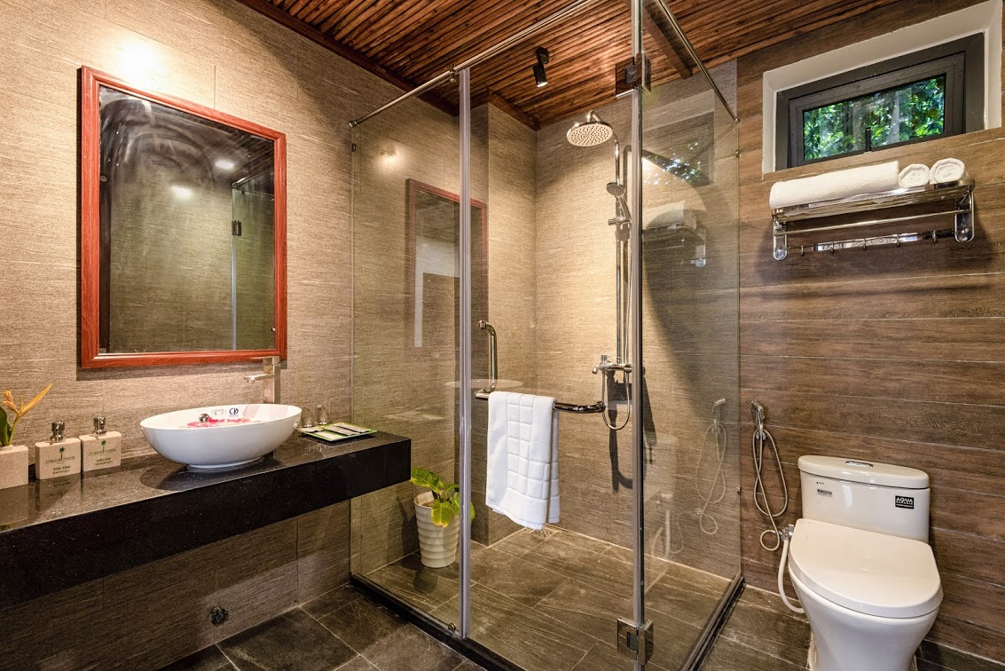 A modern bathroom features a glass-enclosed shower with a rainfall showerhead. A round sink is positioned on a sleek black countertop, and a large mirror reflects the natural light. Complementary toiletries and a potted plant add a touch of warmth to the space.