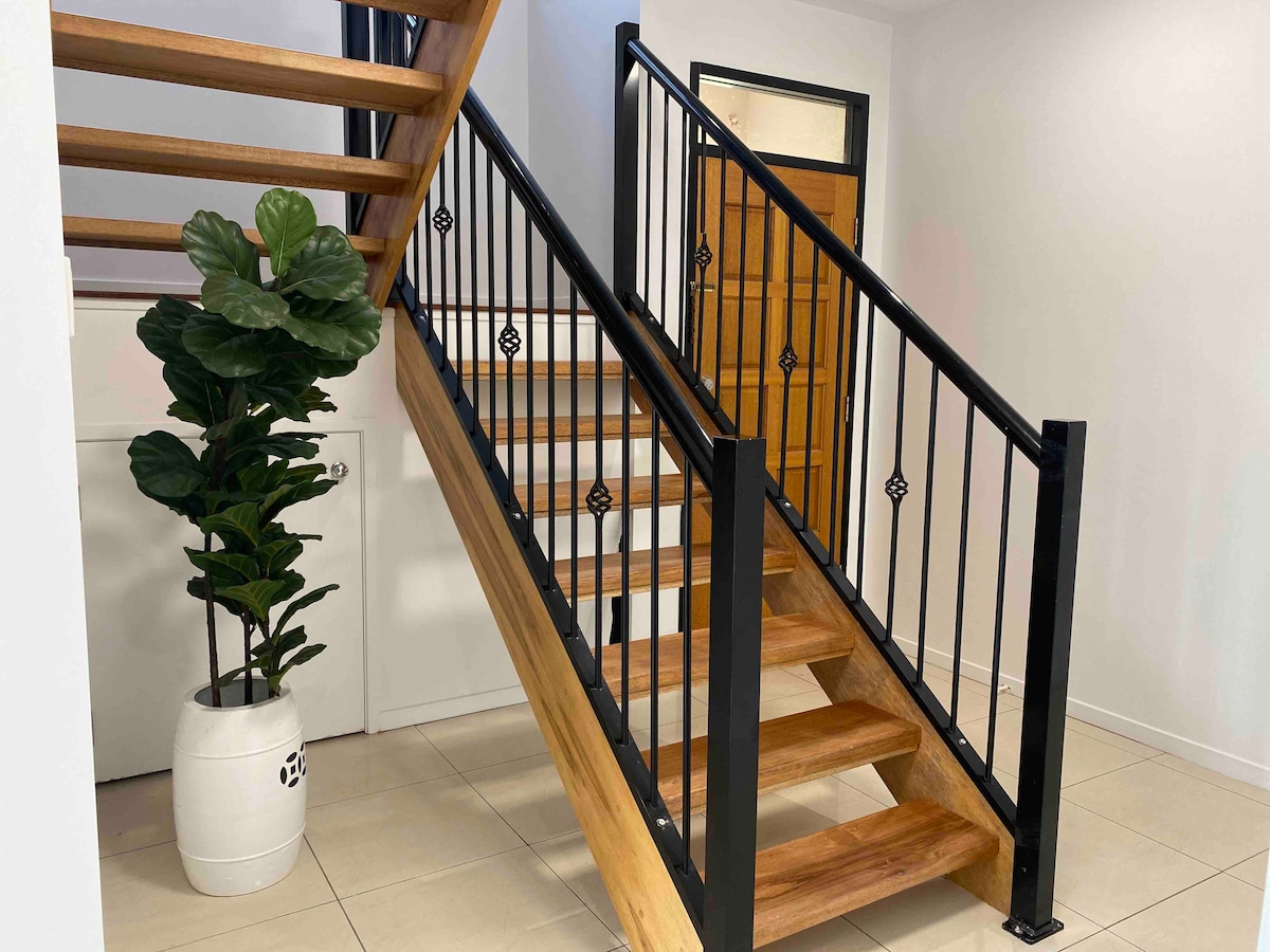 A wooden staircase with black metal railings is featured prominently, leading to an upper level. A large potted plant is positioned beside the stairs, complementing the neutral tiled flooring and white walls. The warm wood tones of the stairs create a welcoming entryway.
