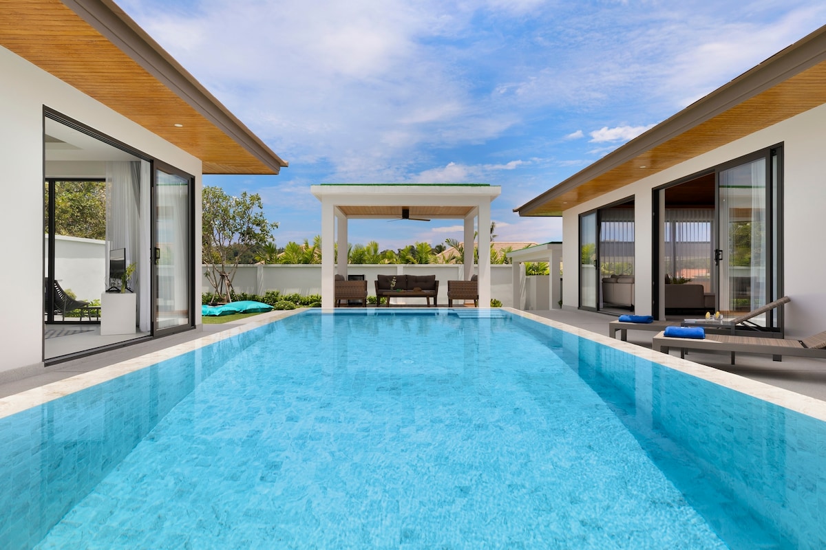 The outdoor area features a sparkling blue swimming pool bordered by white stone tiles. Two pavilions flank the pool, each with large glass doors, providing an airy feel. A shaded seating area is situated at the far end, offering a serene view of the garden.
