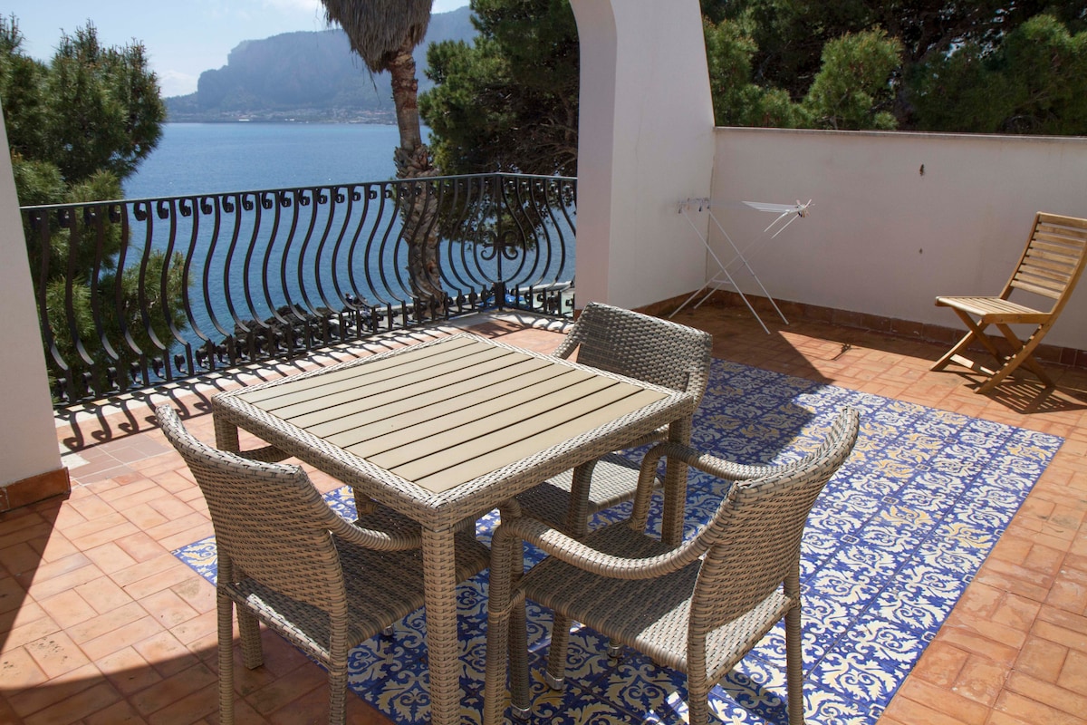 A terrace with a view of the sea features a rectangular table surrounded by four woven chairs. A single wooden lounge chair is positioned nearby, while a clothes drying rack stands against the wall. The floor is tiled with a decorative blue and white pattern.