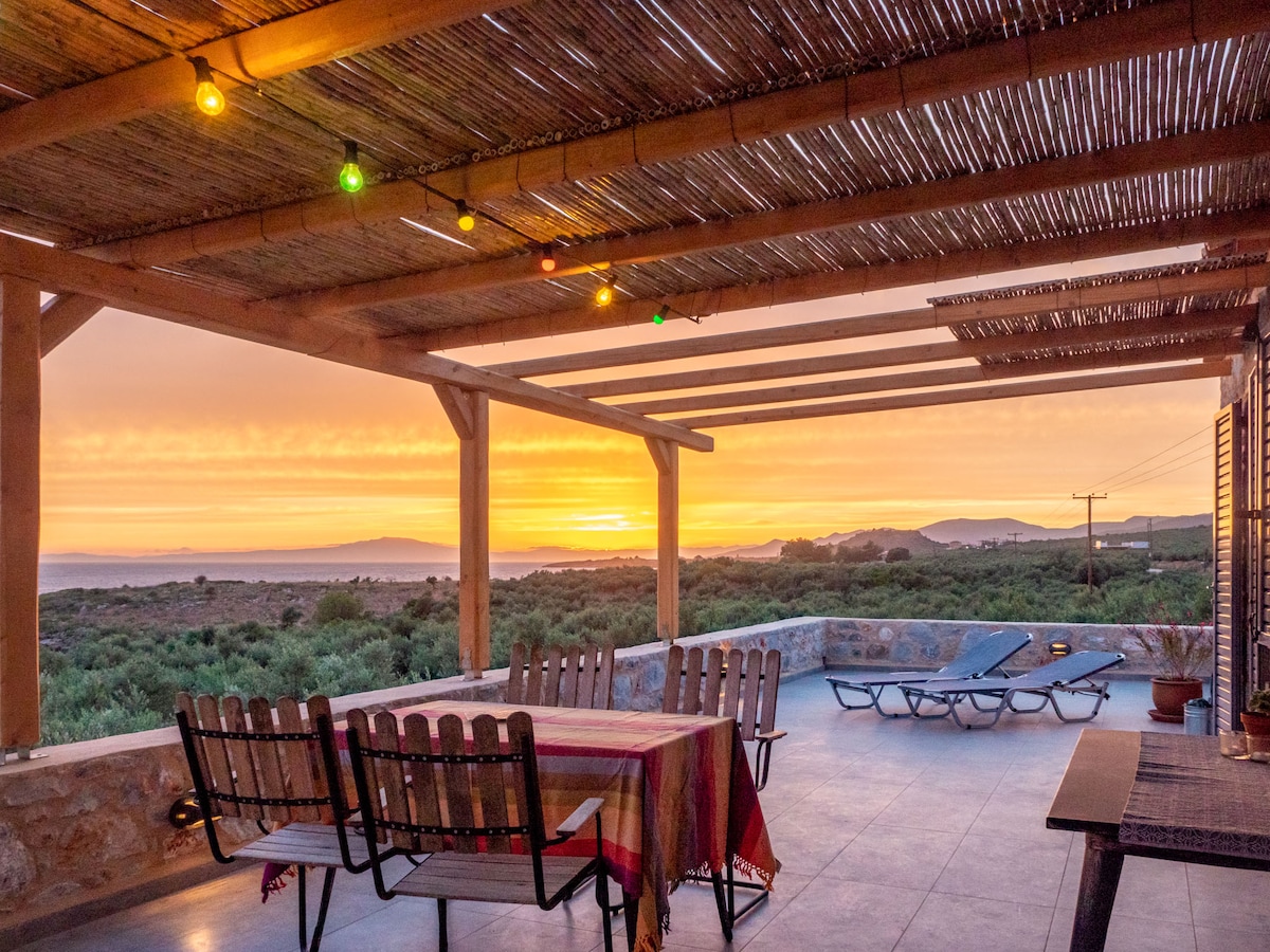 An expansive terrace is featured, adorned with a dining table and several chairs. Colorful lights hang from the wooden overhead structure, illuminating the space as a beautiful sunset casts warm hues over the landscape, with olive groves and distant hills visible in the background.