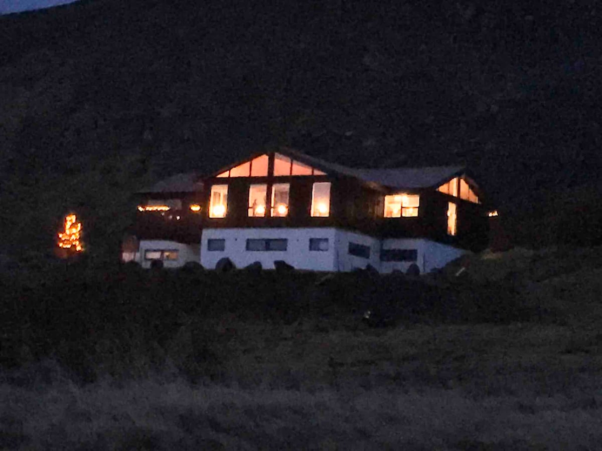 A two-story house is set against a dark mountain backdrop, featuring large windows that glow warmly in the evening light. The structure has a sloped roof and is surrounded by open land, providing an inviting and serene atmosphere.