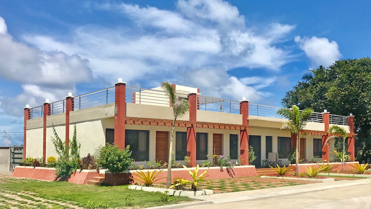A modern one-story building is surrounded by well-maintained landscaping, featuring colorful plants and palm trees. A spacious terrace with railing offers an outdoor area above, while large windows enhance natural light. The structure's clean lines and architecture provide a contemporary aesthetic.