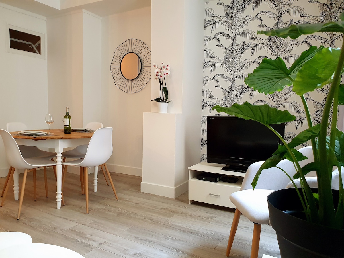 A bright, inviting living space features a round wooden dining table surrounded by four white chairs. A medium-sized television is positioned on a low cabinet. A decorative plant adds a touch of greenery, complemented by a stylish mirror and a wallpaper with a palm leaf design.