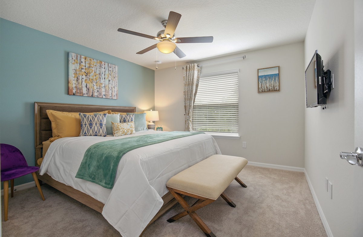 A well-appointed bedroom features a king-sized bed with pastel-colored bedding and an assortment of decorative pillows. A large window, dressed with light curtains, illuminates the space. A stylish bench at the foot of the bed complements the contemporary decor, while a wall-mounted TV adds modern convenience.