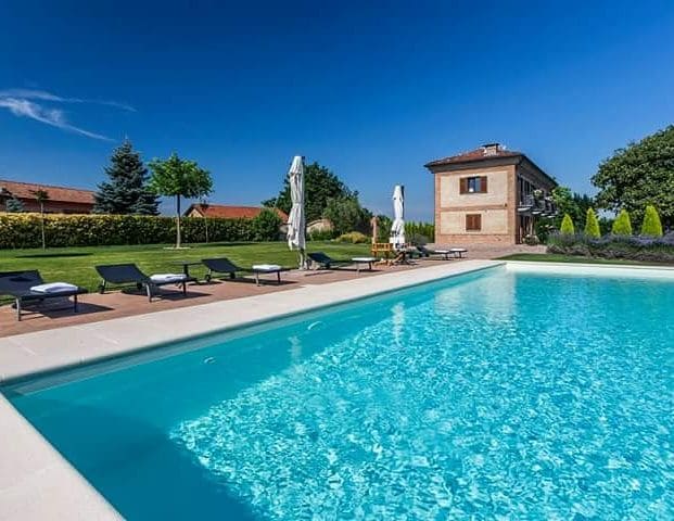 A refreshing infinity pool reflects the clear blue sky, surrounded by lounge chairs and umbrellas. Lush greenery and neatly trimmed hedges frame the pool area, while a charming two-story house is situated in the background, enhancing the serene outdoor environment.