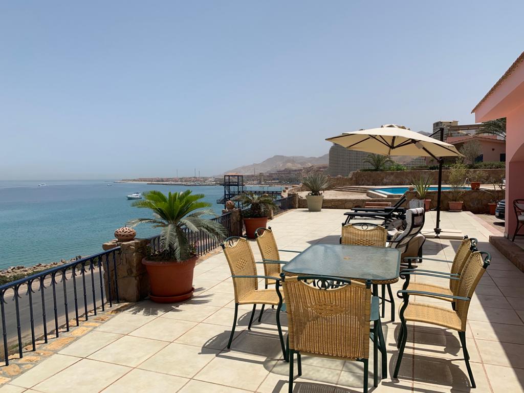 An outdoor terrace features a dining table surrounded by six wicker chairs. Shade is provided by a large umbrella. The area overlooks a tranquil sea, with lounge chairs placed nearby. Coastal plants and a stone railing enhance the serene atmosphere.