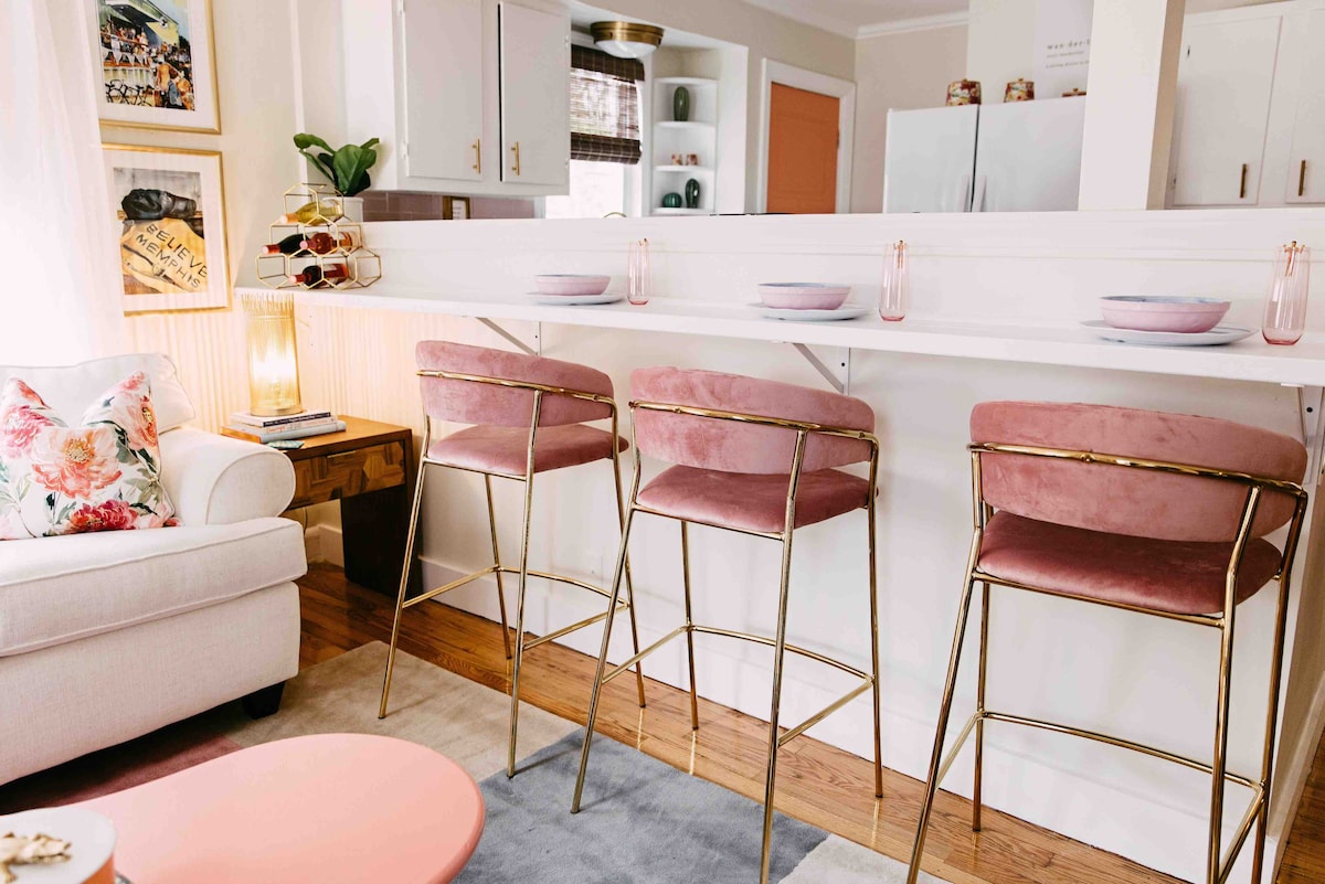 A modern kitchen bar area features three high stools with soft pink upholstery and gold accents. A white countertop is prepared for dining, with pastel plates and elegant drinkware set in place. A cozy sofa with floral cushions is positioned nearby, creating a welcoming space.