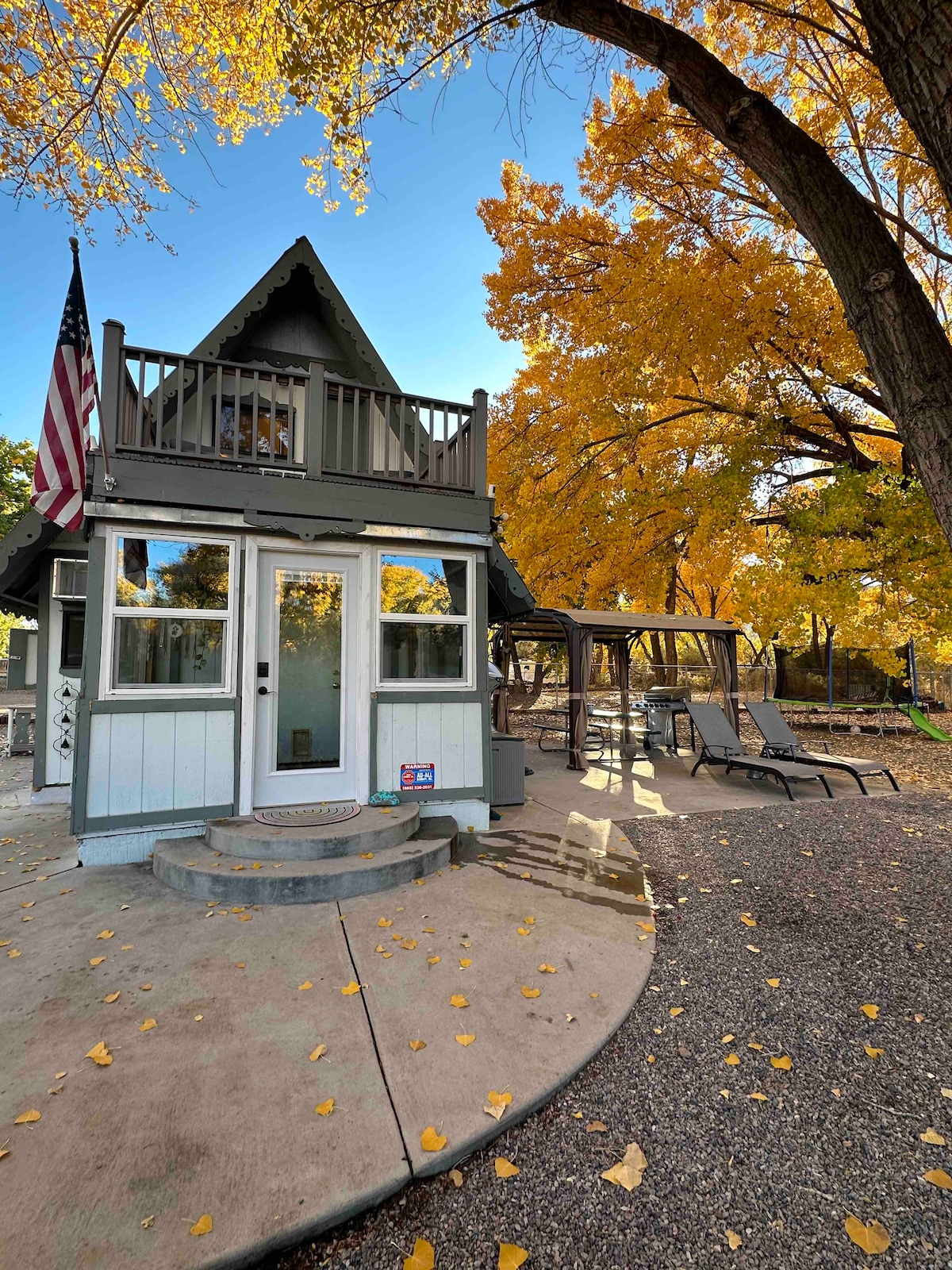 The exterior of the cabin features a charming A-frame design with large windows and a balcony above. Surrounding the cabin are lounge chairs on a gravel patio and a shaded gazebo, framed by vibrant fall foliage from nearby trees.