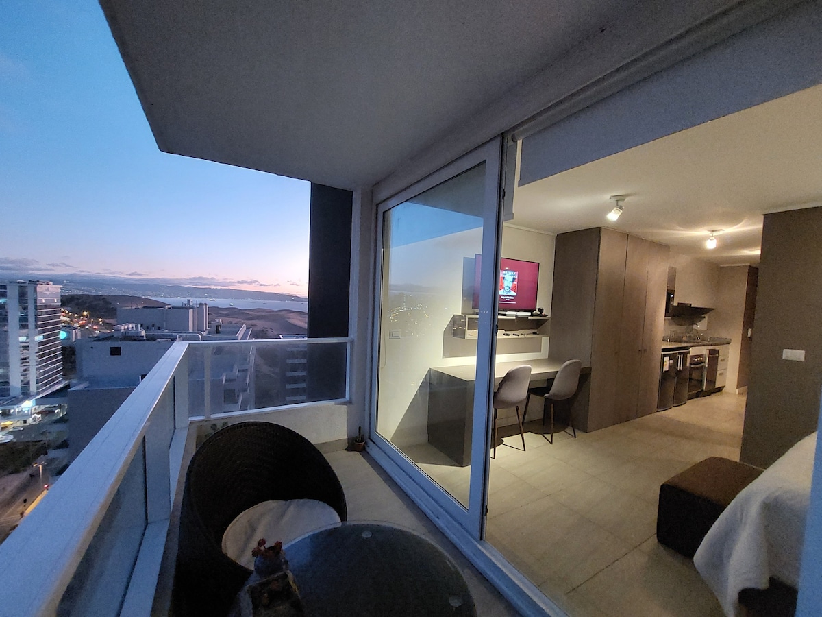 A covered terrace offers a view of the horizon, highlighted by a soft sunset. Seating arrangements include a stylish table and two chairs, while glass doors provide a connection to the indoor living space, which features modern furnishings and a compact kitchen area.