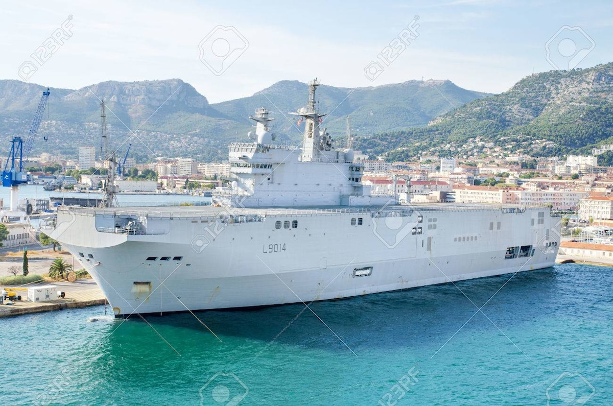 A large naval ship is docked along a coastline, displaying a gray exterior with multiple decks and antennas. The background features mountainous terrain and the waterfront, with buildings integrated into the coastal landscape. A clear sky allows for bright lighting on the scene.
