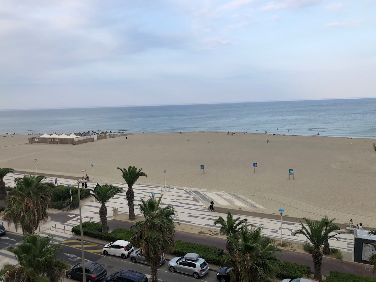A serene view of a sandy beach extends to the horizon, where gentle waves meet the shore. Palm trees line the walkway, providing a tropical touch. Umbrellas are visible in the distance, while a few individuals enjoy the beach area, contributing to a relaxed coastal atmosphere.
