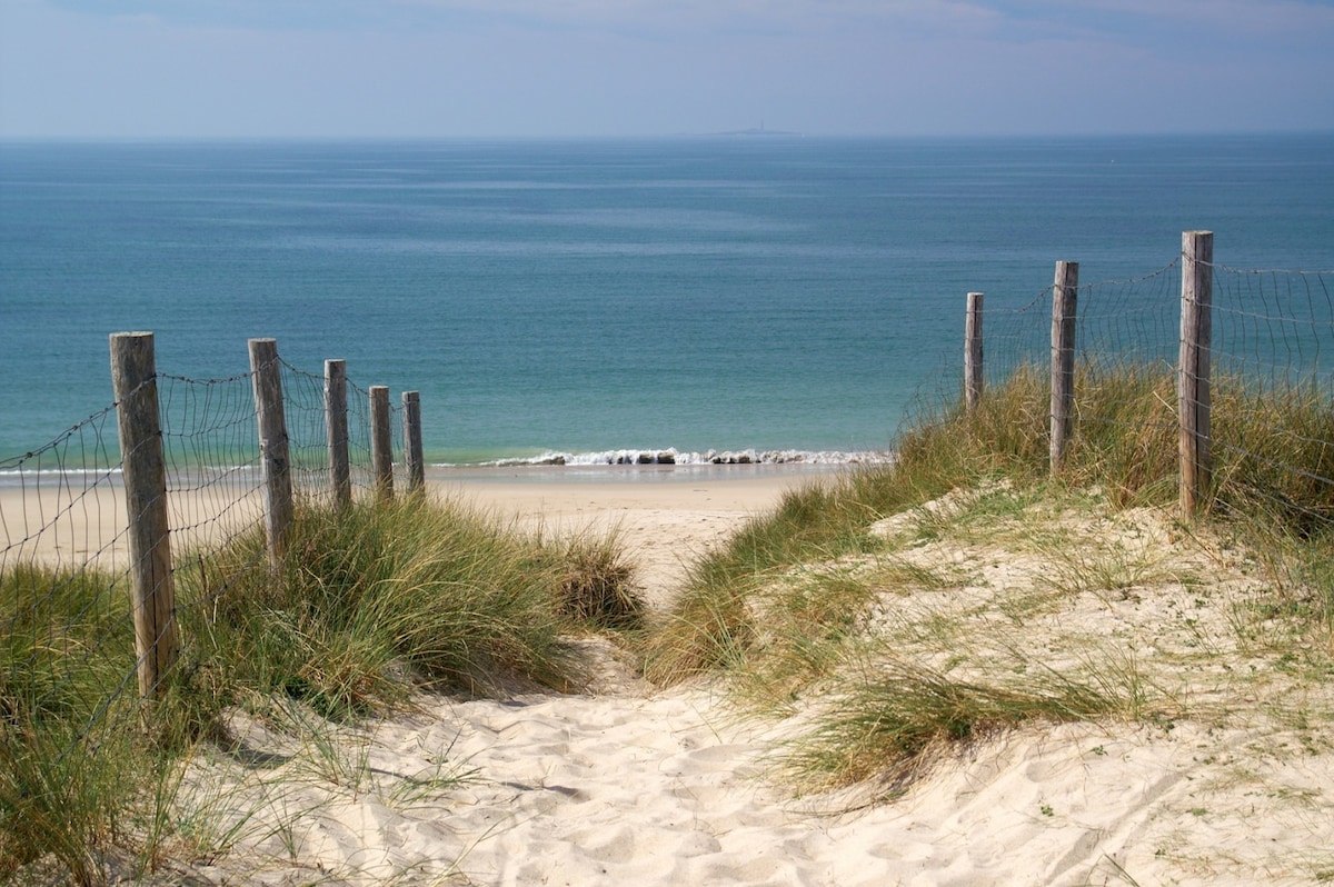 A sandy pathway leads to a serene beach, bordered by grassy dunes and wooden posts with fencing. The tranquil blue sea stretches out in the distance, meeting a gentle shoreline under a clear sky.