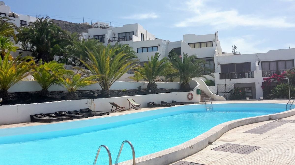 A serene outdoor pool is framed by lush palm trees and landscaped gardens, with lounge chairs positioned nearby. White buildings with large windows are visible in the background, reflecting a calm atmosphere under a clear blue sky.