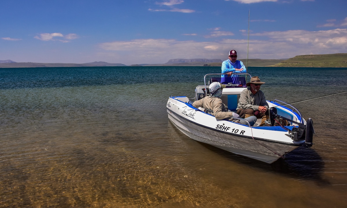 A fishing boat rests on clear waters, surrounded by a scenic landscape. Two individuals are seated, engaged in conversation. The distant shore features rolling hills under a bright sky with scattered clouds, creating a serene environment ideal for outdoor activities.