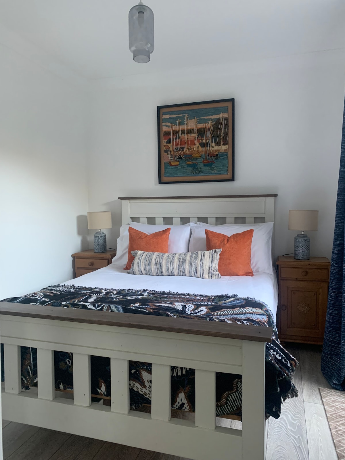A well-lit bedroom features a spacious bed with a dark blanket and decorative cushions in orange and blue. Two wooden nightstands with lamps are positioned on either side of the bed. A nautical-themed painting hangs above the headboard, adding character to the room.