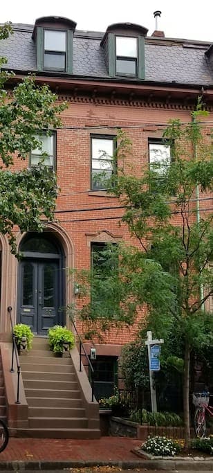 A historic townhouse features a striking red brick exterior, accented by distinctively styled windows. A set of steps leads to a prominent double door entrance, flanked by planters with greenery. Trees and shrubs enhance the welcoming street view, inviting exploration of the neighborhood.