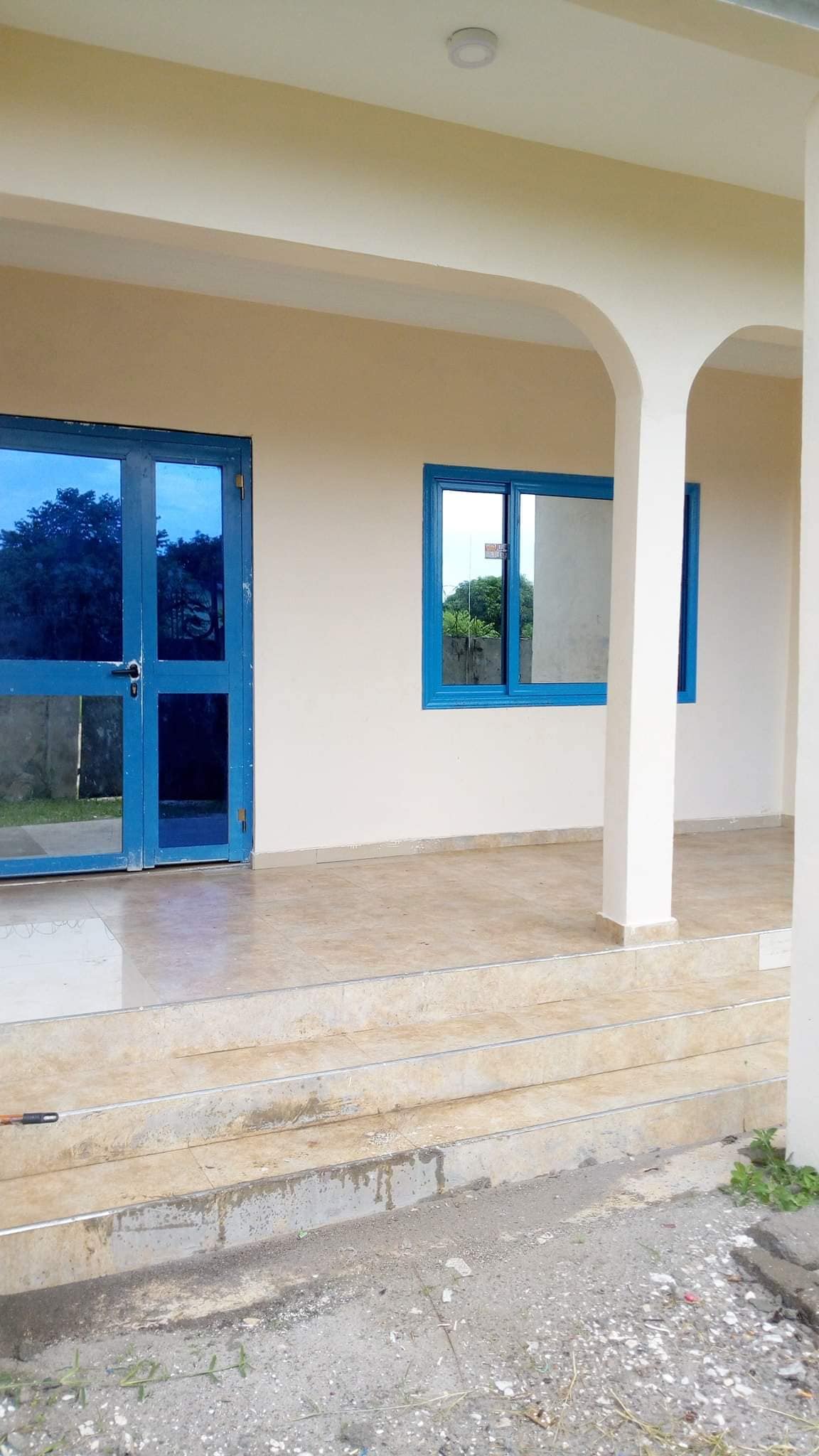 A welcoming entrance is presented with a blue front door and matching window frames. The floor is finished in smooth, light-colored tiles, leading up to a covered porch. Clean lines and soft colors create a calm and inviting space.