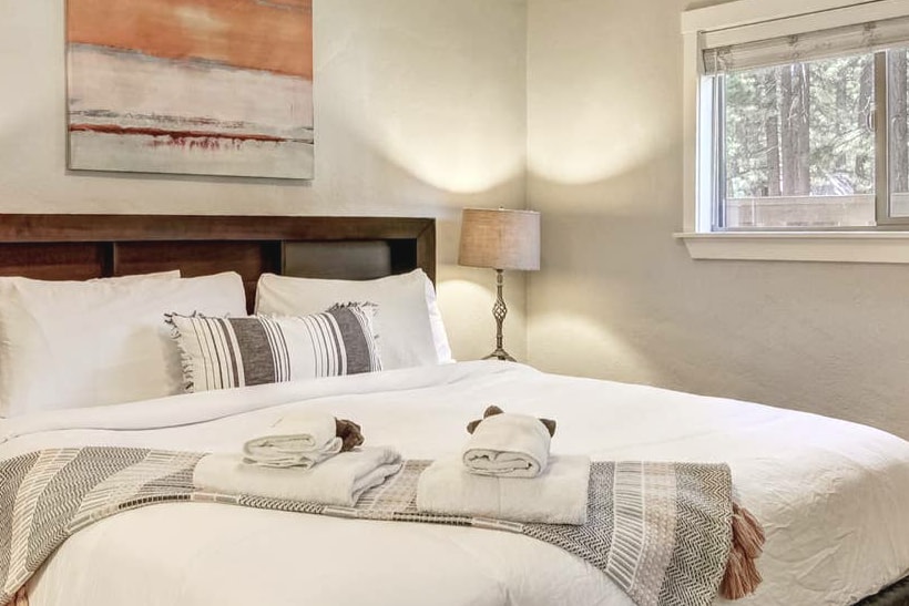 A softly lit bedroom features a spacious bed adorned with neutral linens and decorative pillows. A light-colored painting hangs above the headboard, while a bedside lamp provides additional warmth. Natural light enters through a large window dressed with minimalistic blinds.