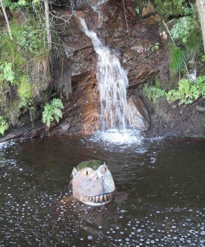 A whimsical sculpture resembling a creature emerges from dark waters, surrounded by lush greenery. A gentle waterfall cascades down a rocky surface in the background, creating ripples in the water below. The scene conveys a serene connection to nature.