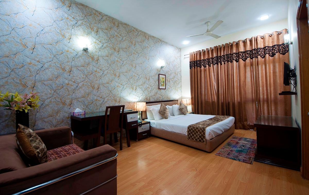 A spacious bedroom is highlighted by decorative wallpaper and soft lighting. A large bed is positioned against a wall, accompanied by nightstands. A plush sofa and a wooden dining set are visible, with rich brown curtains framing the window, allowing for a warm and inviting feel.