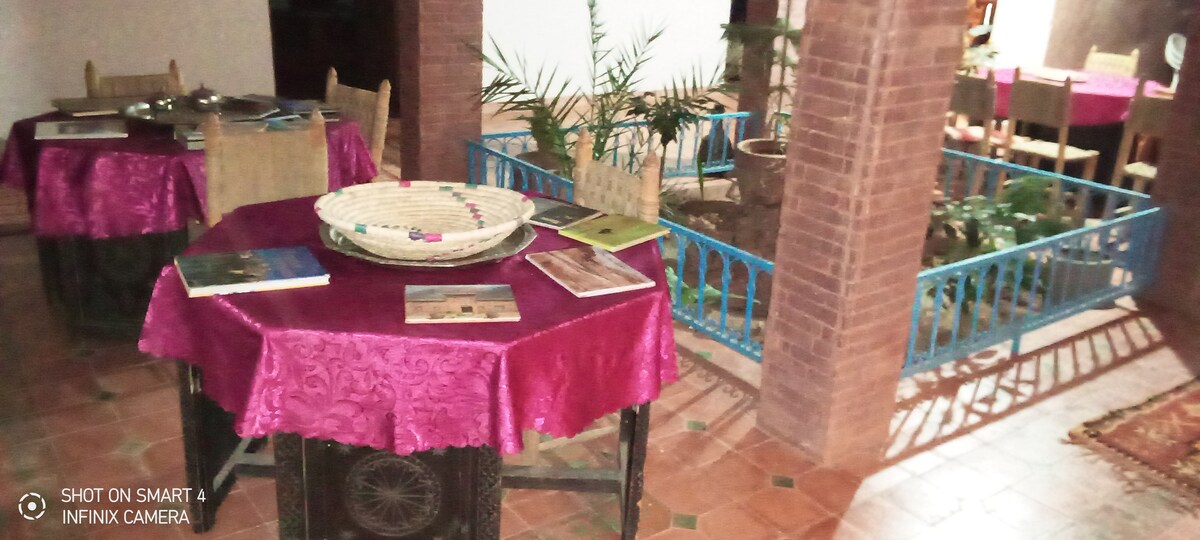 Dining tables are arranged with vibrant pink tablecloths and decorative items, creating a warm dining area. Potted plants are visible in the background, alongside a low wooden railing separating the space from an indoor garden, contributing to the serene ambience.