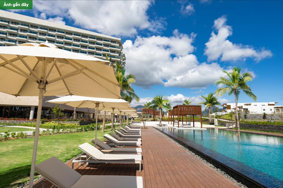 A wide outdoor area is depicted with lounge chairs lined along a wooden deck, facing a swimming pool. Umbrellas provide shade, and palm trees frame the scene. A well-maintained garden area is shown in the background, with clear blue skies and scattered clouds above.