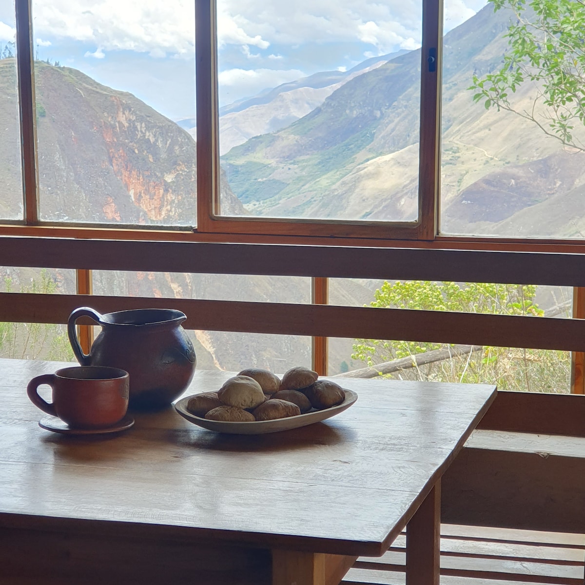 A wooden table is set with a plate of baked goods and a beverage cup beside a clay pitcher. Outside, expansive mountain views are framed by large windows, bringing in natural light and highlighting the serene landscape.