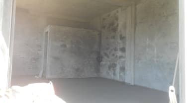 An unfinished room is presented with raw concrete walls and an expansive, unobstructed floor area. A small, partitioned space is visible towards the back, suggesting potential for customization or privacy.