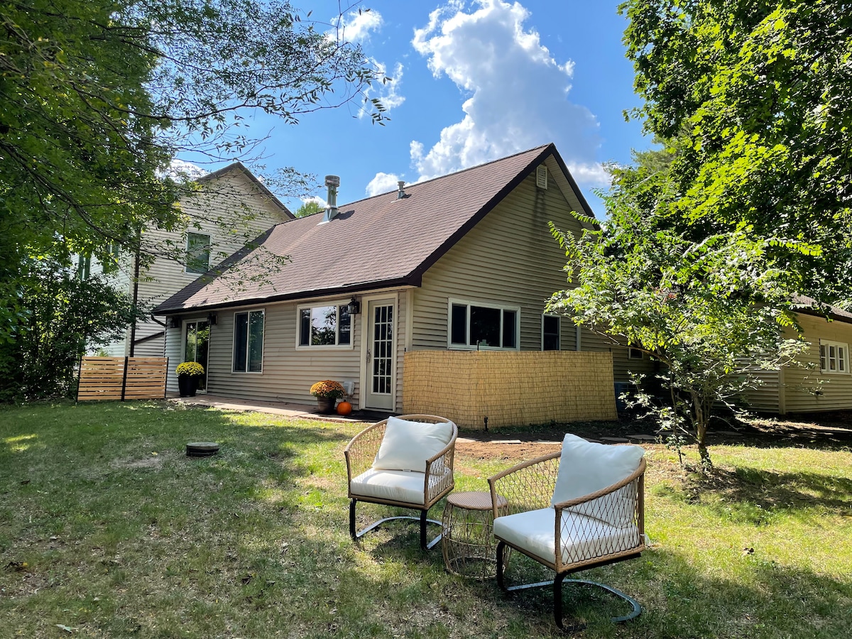 A view of the exterior reveals a cozy single-story home nestled among trees. Two chairs with a small table are positioned on green grass, inviting outdoor relaxation on a sunny day. The structure features a gently sloped roof and large windows, enhancing natural light.