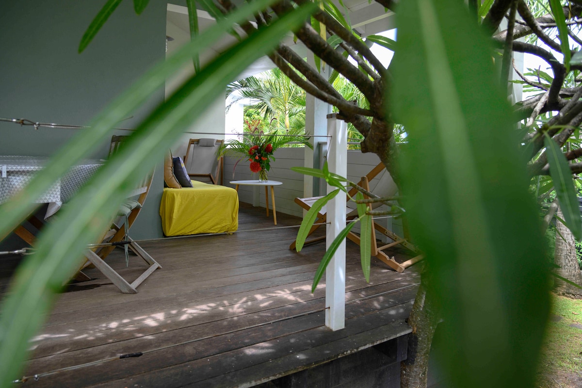A shaded terrace is adorned with wooden flooring and surrounded by lush greenery. Two angled chairs and a small table are positioned, with a vibrant floral arrangement enhancing the setting. The area provides a peaceful retreat for relaxation.