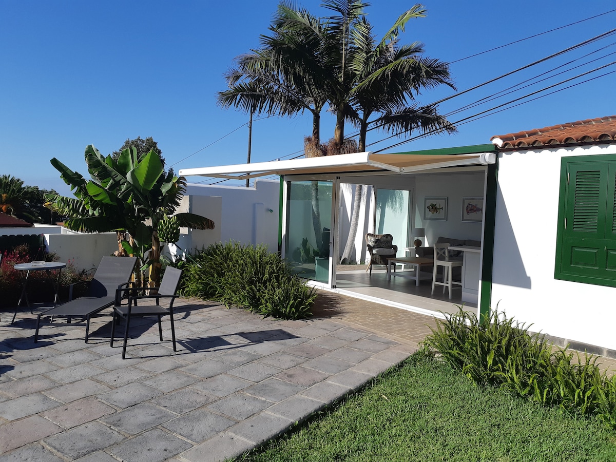 A modern cottage is presented with sliding glass doors opening to a patio area. Tropical plants, including a tall palm, frame the space. Outdoor seating is set up with chairs and a small table on a stone patio, surrounded by landscaped greenery.