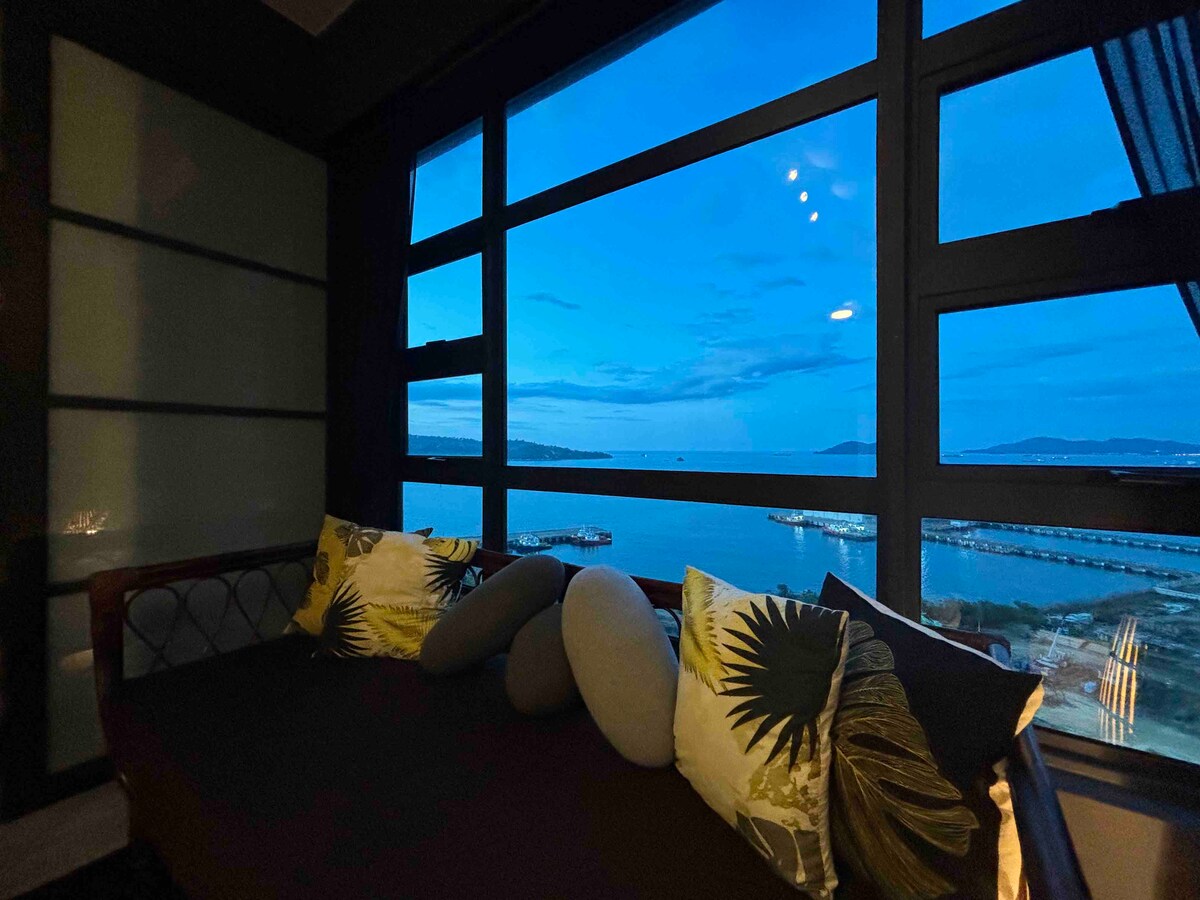 A cozy lounging area features a large window offering a panoramic view of the water at twilight. Decorative pillows add comfort to the daybed, while soft illumination provides a gentle ambiance. Various shades of blue dominate the sky and water beyond the window.