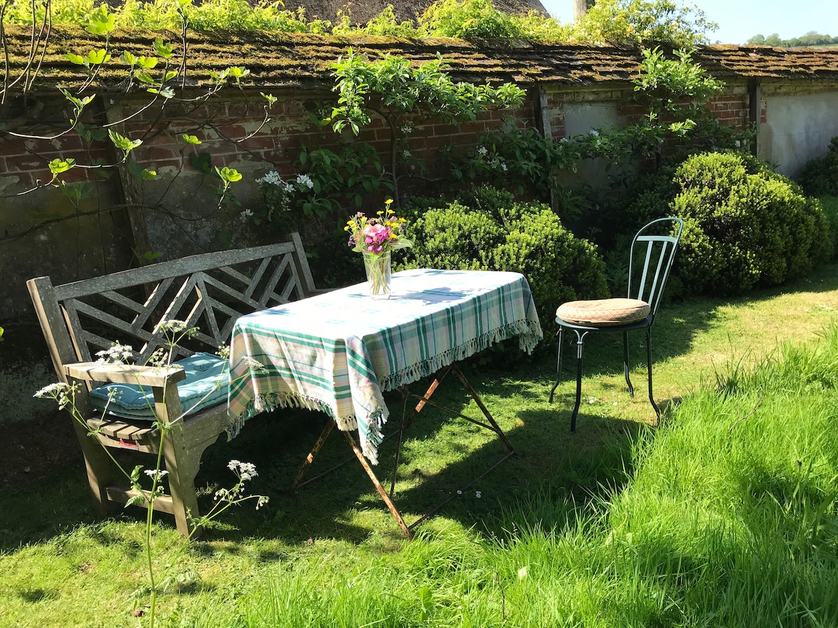 An outdoor garden space is shown, featuring a rustic wooden bench alongside a small table draped with a checkered cloth. A vase of flowers sits on the table, surrounded by neatly trimmed shrubs and foliage in a sunny, green setting.