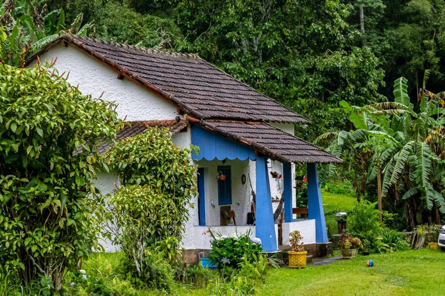 A charming white house features blue accents and a covered porch. Surrounding greenery includes lush plants and small trees, adding to the natural setting. The gentle slope of the landscape enhances the rustic appeal of the home, providing a serene view of the countryside.