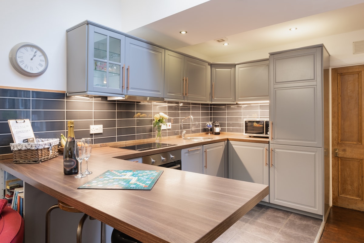 The modern kitchen is fitted with gray cabinetry and a brown countertop. A wine bottle and glasses are neatly placed on the counter, alongside a colorful board game. Ambient lighting casts a gentle glow, enhancing the inviting layout with appliances and space for socializing.