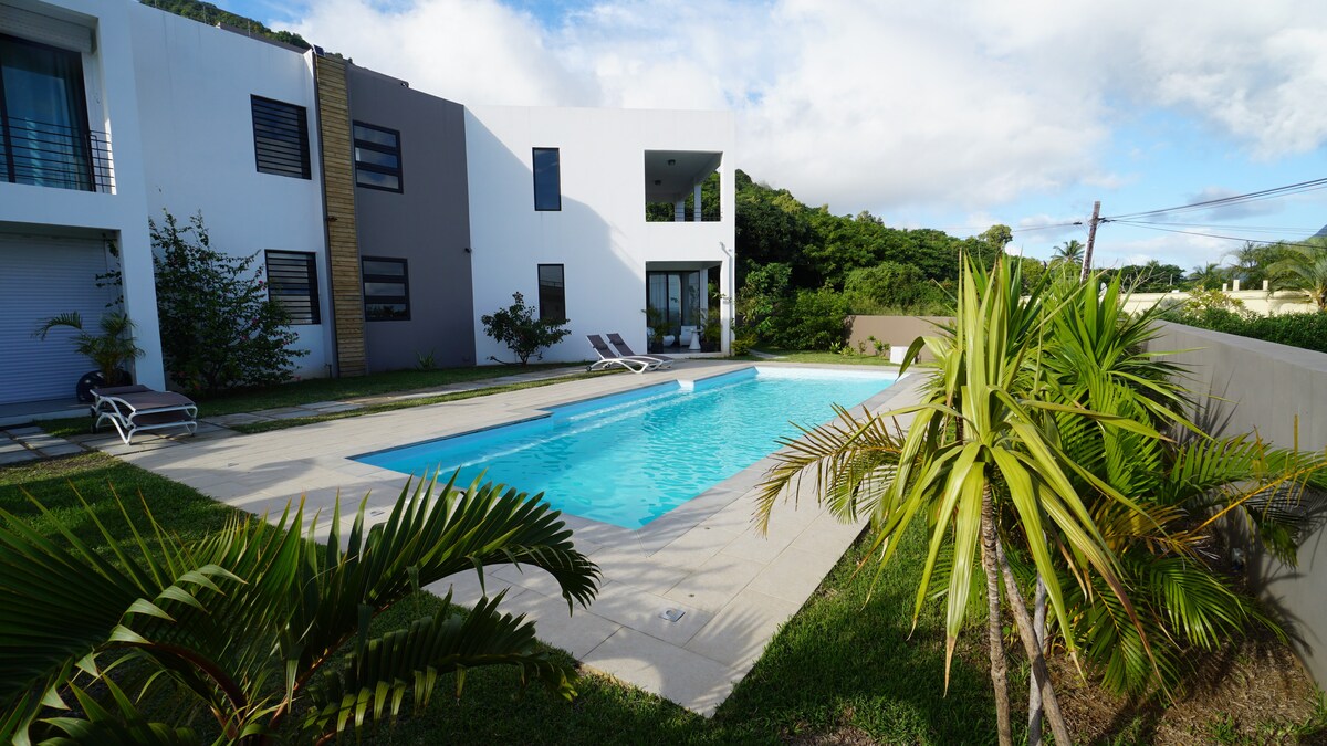 A spacious outdoor area features a rectangular swimming pool surrounded by a concrete patio. Lounge chairs are positioned beside the pool, with greenery and palm plants creating a natural border. The modern building structure is visible in the background, with large windows reflecting the sky.