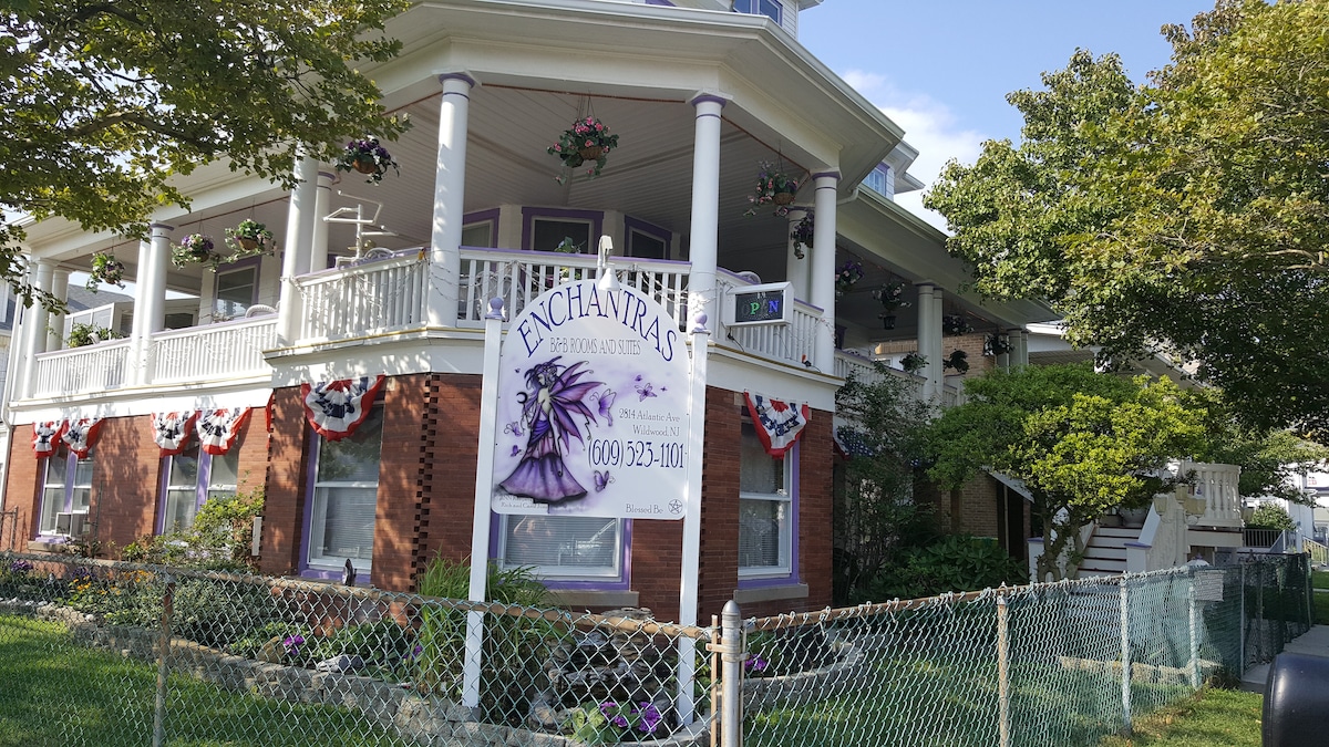 The exterior of a charming Victorian home is displayed, featuring a welcoming wrap-around porch adorned with hanging flower baskets. A sign with the name 'Enchantress' is prominently positioned. Lush greenery and colorful flower beds complement the inviting atmosphere of the property.