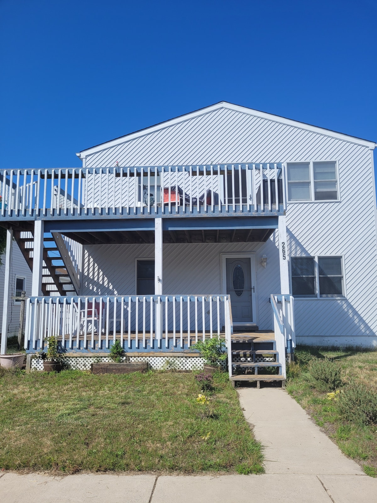 A two-story home is presented with a bright white exterior and a covered front porch. The porch features railings and a set of stairs leading to the upper level. A door is centered below large windows, framed by simple landscaping on the grassy front yard.