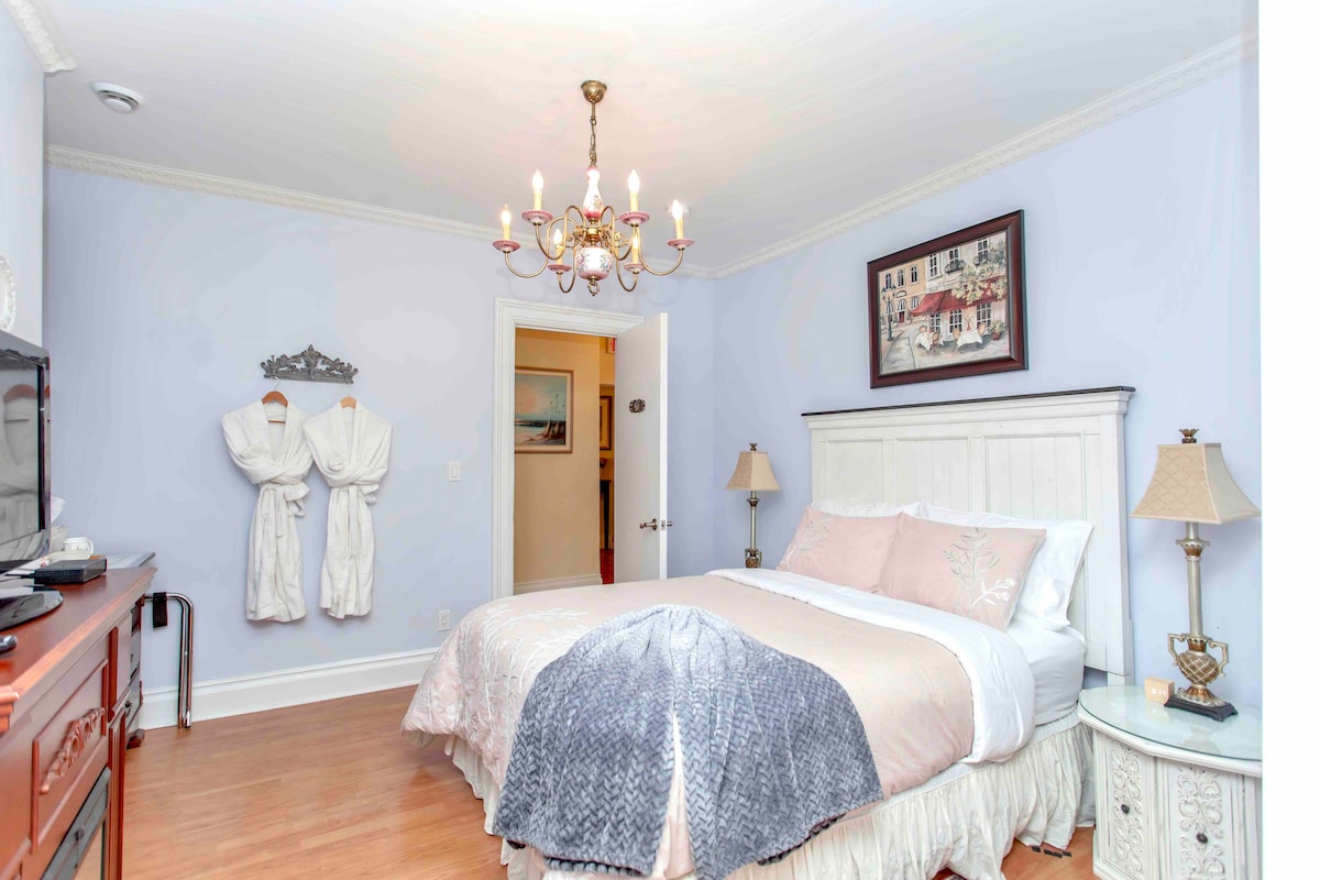 The guest room features a spacious bed dressed in soft linens and pillows, complemented by a light-colored decorative throw. A chandelier illuminates the room, while two bathrobes hang neatly on the wall. A television and bedside lamps are situated on either side of the bed.