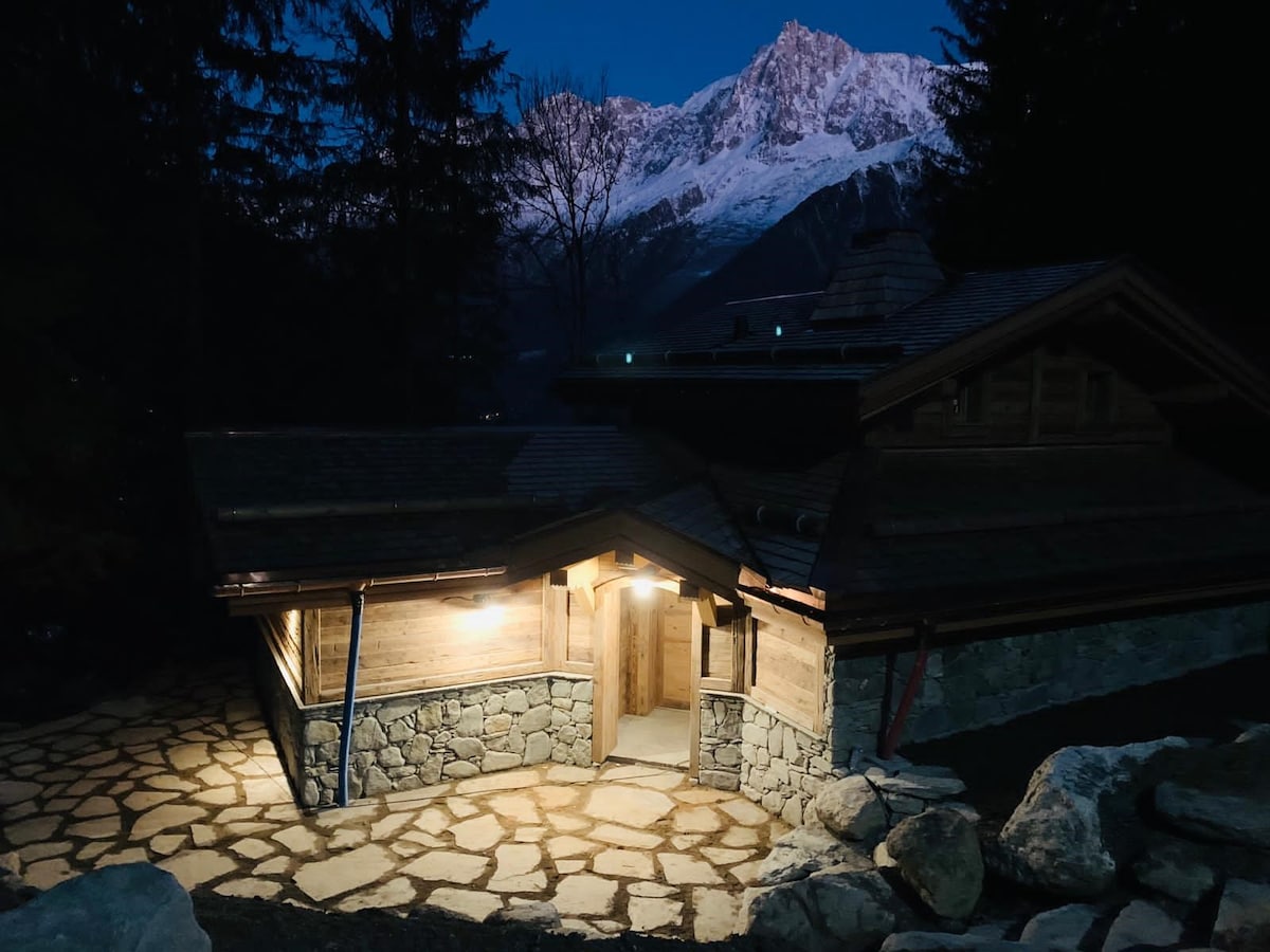 The chalet is illuminated at night, featuring a stone pathway leading to a wooden entrance. Surrounding trees are visible, with the majestic snow-capped mountains providing a dramatic backdrop against the evening sky.