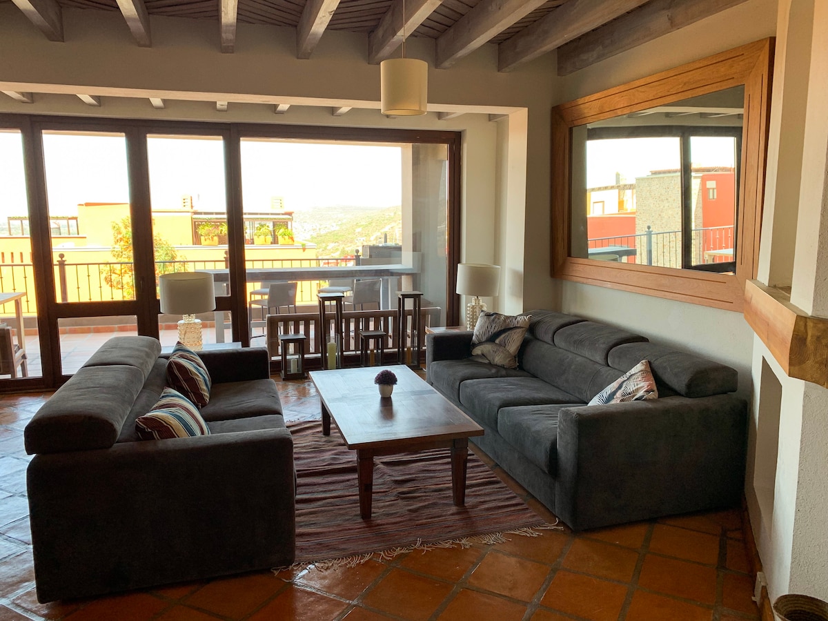 A spacious living area features large windows that allow for abundant natural light and offer scenic views. Two dark sofas are positioned around a central wooden coffee table, with a decorative rug adding warmth to the tiled floor. A large mirror enhances the open ambiance.