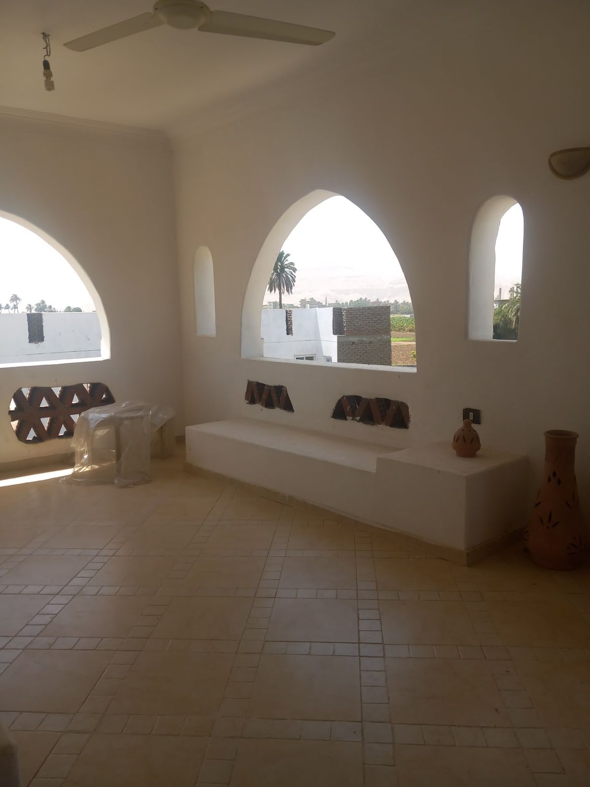 An open terrace area showcases arched windows that provide views of the surrounding landscape. A white bench is integrated into the design, complemented by decorative pottery positioned thoughtfully around the space. Natural light illuminates the tiled floor, enhancing the airy ambiance.