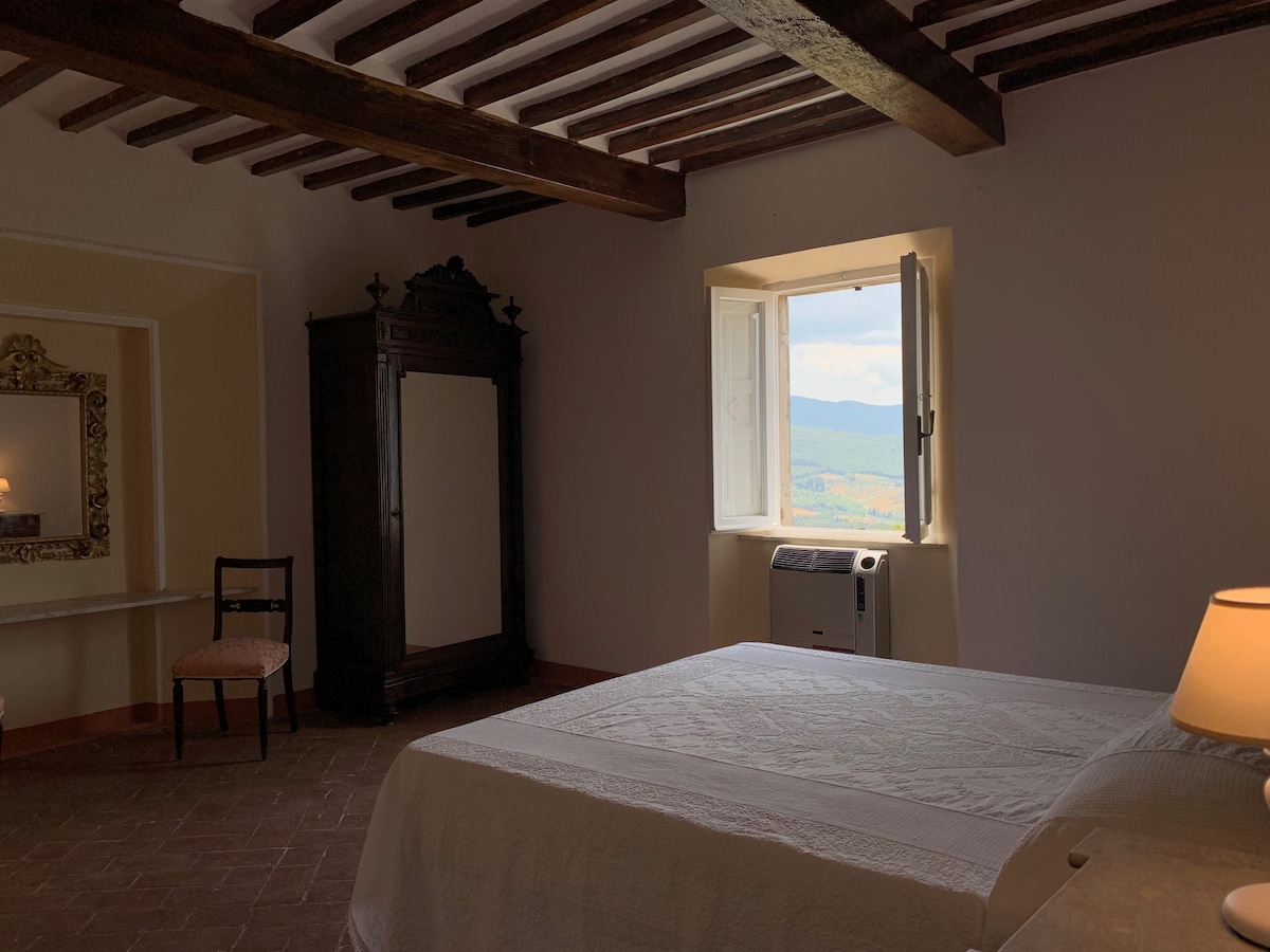 A spacious room features a large bed with a textured white coverlet and natural light coming through an opening window. An antique wardrobe stands against the wall, and a simple wooden chair is positioned nearby. A vanity area with a mirror completes the calming ambiance.
