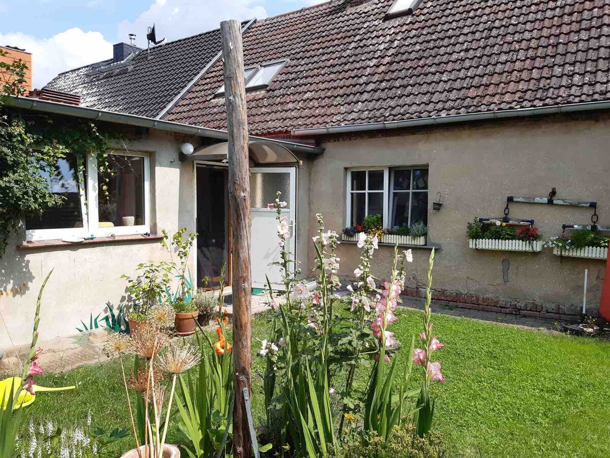 A charming garden area is showcased with vibrant flowers and greenery. The house, featuring a rustic facade, is visible in the background, along with an entrance framed by a simple structure. Natural light enhances the inviting outdoor space.