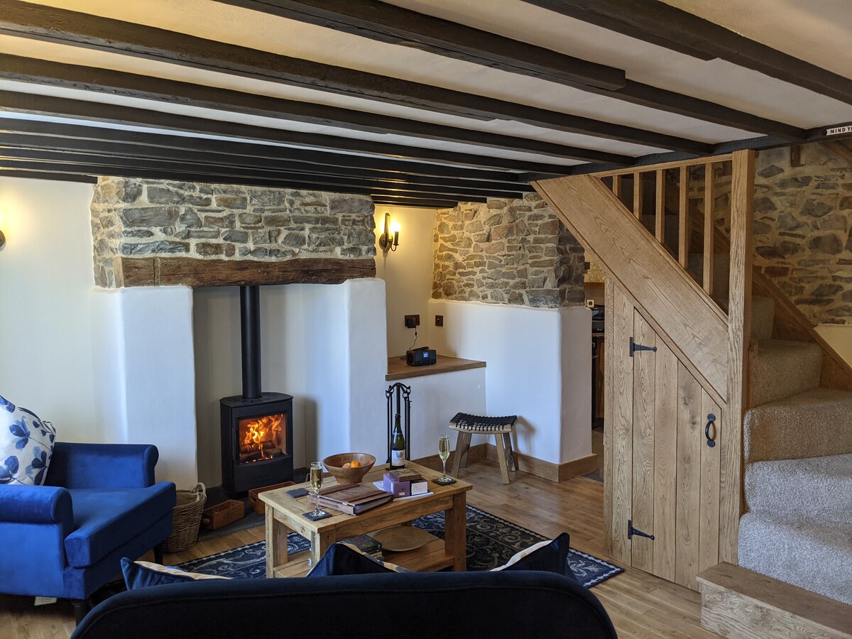 A cozy sitting area features a blue armchair and a wooden coffee table near a stone fireplace with a wood-burning stove. Exposed wooden beams and warm lighting create a welcoming ambiance, while stairs lead to an upper level, enhancing the cottage's rustic charm.