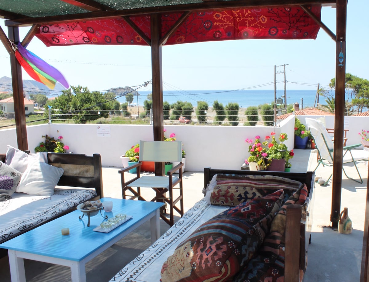 A large roof terrace is presented with shaded seating areas featuring comfortable cushions and decorative throws. Colorful potted flowers enhance the space, while a view of the nearby beach and ocean can be seen in the background. A rainbow flag adds a vibrant touch.
