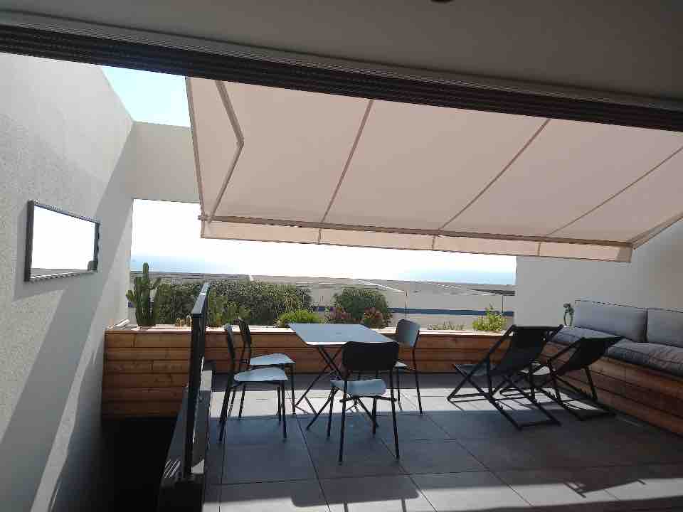 A spacious terrace features a shaded area with a surface covered in gray tiles. A rectangular table and several chairs are positioned on one side, while a cushioned seating area runs along a wall. The ocean view is visible in the distance.